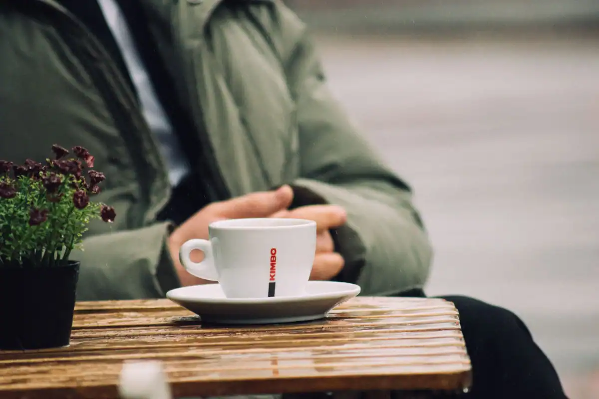 Ceașcă de cafea pe o masă de lemn, cu o persoană așezată în aer liber
