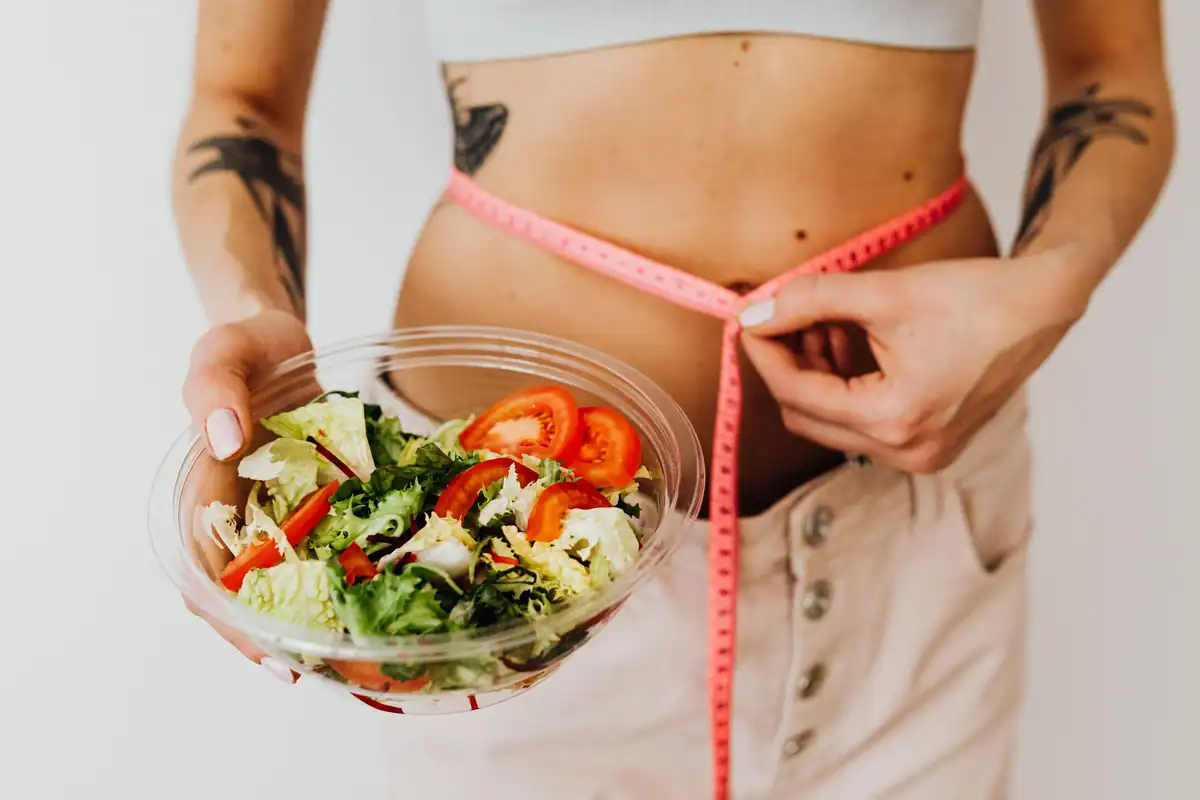 Femeie care ține o salată de legume și își măsoară talia cu un centimetru